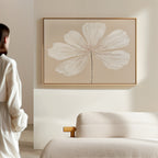 A woman in a white outfit admires the Ivory Bloom Canvas Art - Horizontal, featuring a single white flower on a thin stem, displayed above a beige sofa in a minimalist room with light, neutral décor.