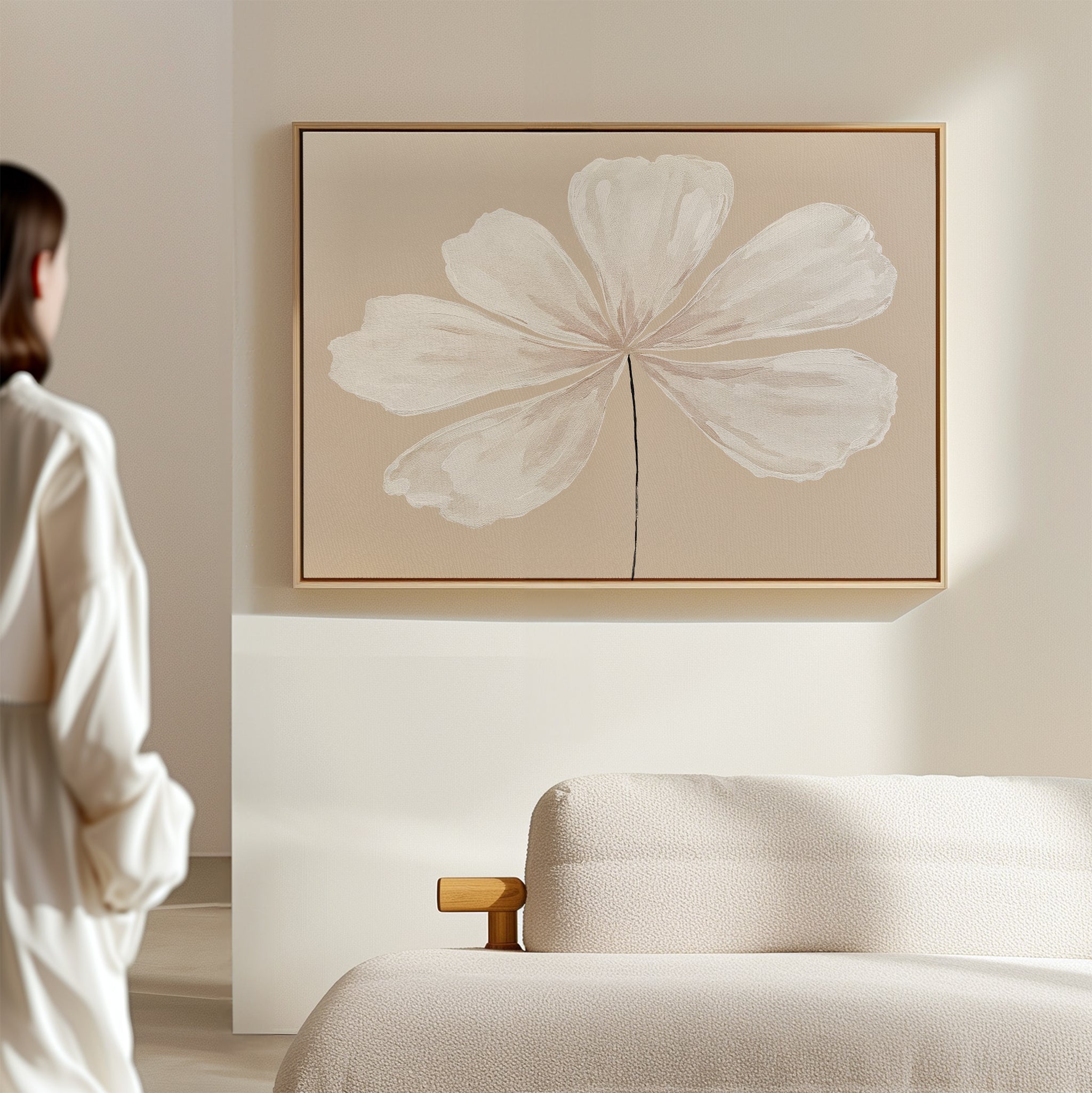 A woman in a white outfit admires the Ivory Bloom Canvas Art - Horizontal, featuring a single white flower on a thin stem, displayed above a beige sofa in a minimalist room with light, neutral décor.