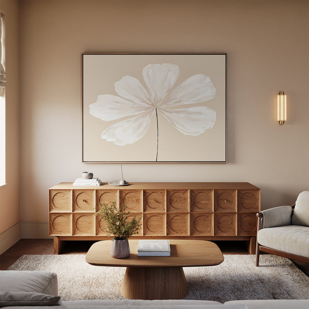 A modern living room with neutral tones showcases the Ivory Bloom Canvas Art - Horizontal above a wooden credenza, paired with a light wood coffee table, books, a beige sofa, and a wall sconce for a calm, minimalist vibe.