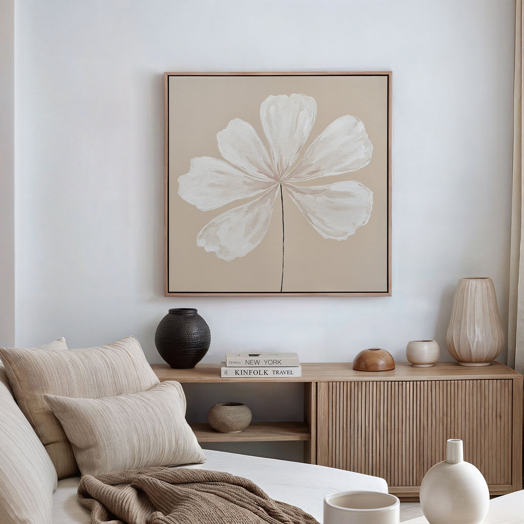 A minimalist living room features a beige sofa, neutral pillows, wooden sideboard with vases and books, and the Ivory Bloom Canvas Art - Square—a striking floral piece with white petals on beige—perfect for neutral home decor.