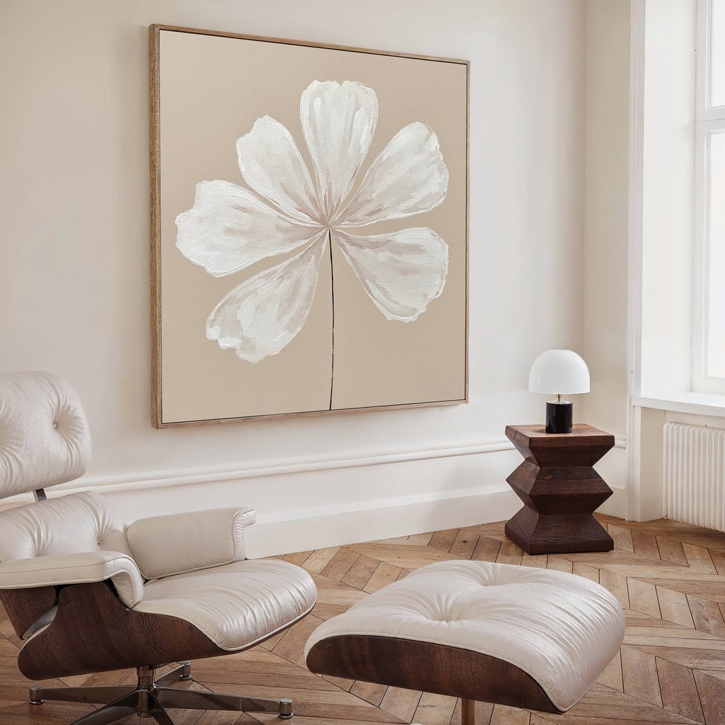 A minimalist living room showcases the Ivory Bloom Canvas Art - Square, a modern cream lounge chair with ottoman, a sculpted wooden side table, and a small white lamp by a tall window for neutral home decor.