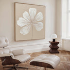 A minimalist living room showcases the Ivory Bloom Canvas Art - Square, a modern cream lounge chair with ottoman, a sculpted wooden side table, and a small white lamp by a tall window for neutral home decor.