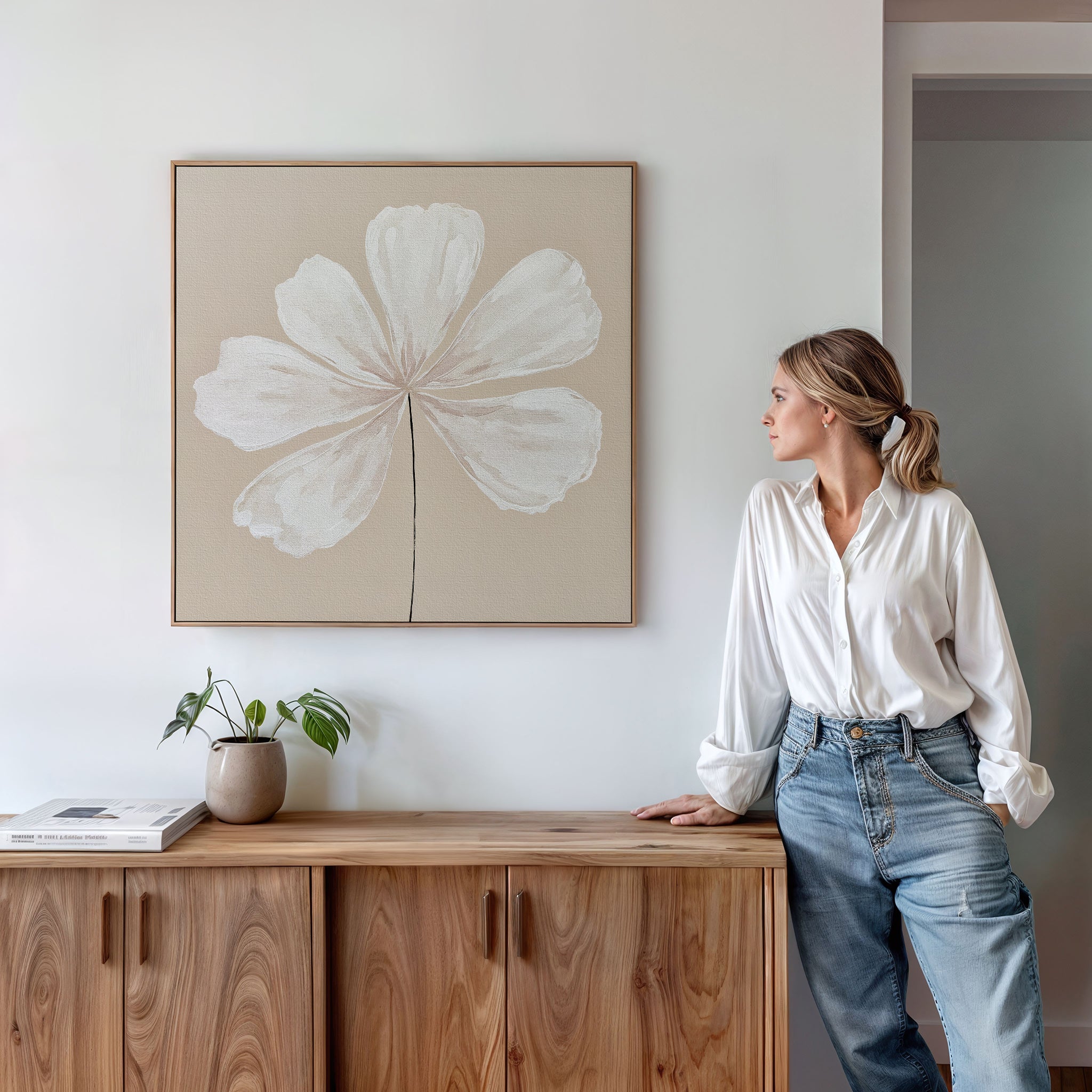 A woman in a white blouse and jeans leans against a wooden cabinet, admiring the Ivory Bloom Canvas Art - Square featuring a large white flower on the wall. A potted plant and books complement the neutral home decor atop the cabinet.