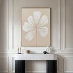 A minimalist interior showcases the Ivory Bloom Canvas Art - Vertical, a large beige botanical artwork of a white flower, mounted above a marble console table adorned with modern candles and geometric vases against an elegant paneled wall.