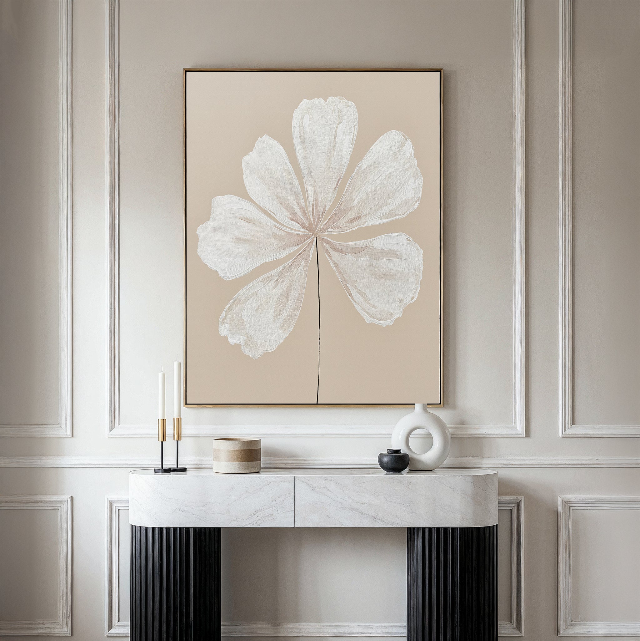 A minimalist interior showcases the Ivory Bloom Canvas Art - Vertical, a large beige botanical artwork of a white flower, mounted above a marble console table adorned with modern candles and geometric vases against an elegant paneled wall.