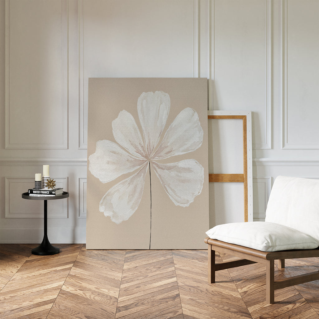 A minimalist room with herringbone wood floors features the Ivory Bloom Canvas Art - Vertical of a white flower against a white paneled wall, alongside a cushioned chair and round table with candles and books—ideal for neutral home decor.