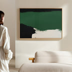 A person in a white shirt stands in a sunlit, minimal room, admiring the Jade Depths Canvas Art - Horizontal with green, black, and white sections displayed above a beige sofa.