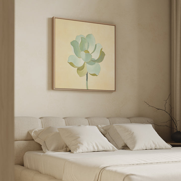 A neatly made bed with beige bedding and multiple pillows sits beneath the Jade Reflection Canvas Art - Square, featuring a green botanical design, on a cream wall in a softly lit, minimalist bedroom.