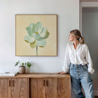 A woman in a white blouse and jeans leans against a wooden cabinet, gazing at the Jade Reflection Canvas Art - Square, a green and yellow botanical print hanging on the white wall above her. A potted plant and books rest on the cabinet.