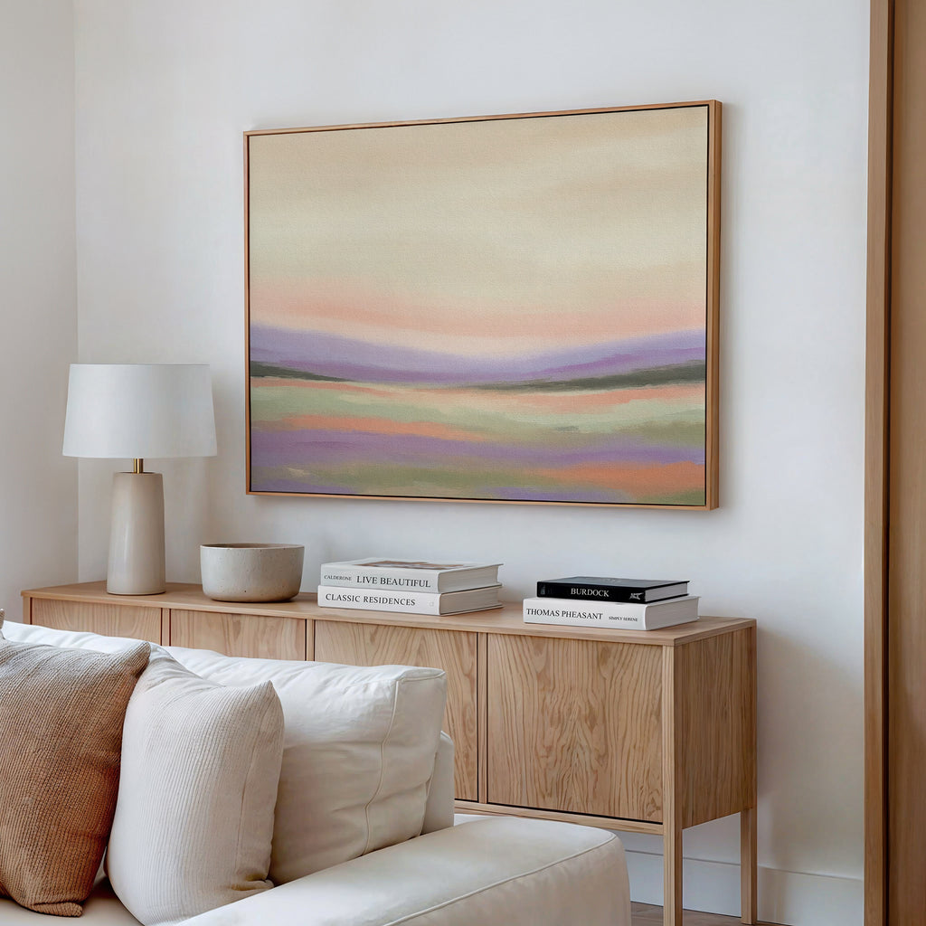 A modern living room features the Lavender Valley Canvas Art - Horizontal, a neutral-toned abstract landscape, hung above a cream-colored sofa on a crisp white wall, with a wooden sideboard, stacked books, and a white table lamp nearby.
