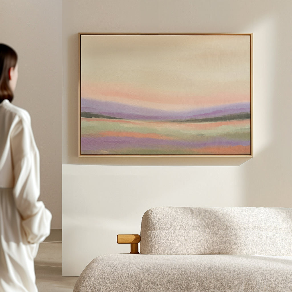 A person in a white robe stands in a minimalist room, gazing at the Lavender Valley Canvas Art - Horizontal, a pastel abstract landscape painting, displayed on a beige wall above a light-colored sofa.