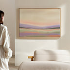 A person in a white robe stands in a minimalist room, gazing at the Lavender Valley Canvas Art - Horizontal, a pastel abstract landscape painting, displayed on a beige wall above a light-colored sofa.