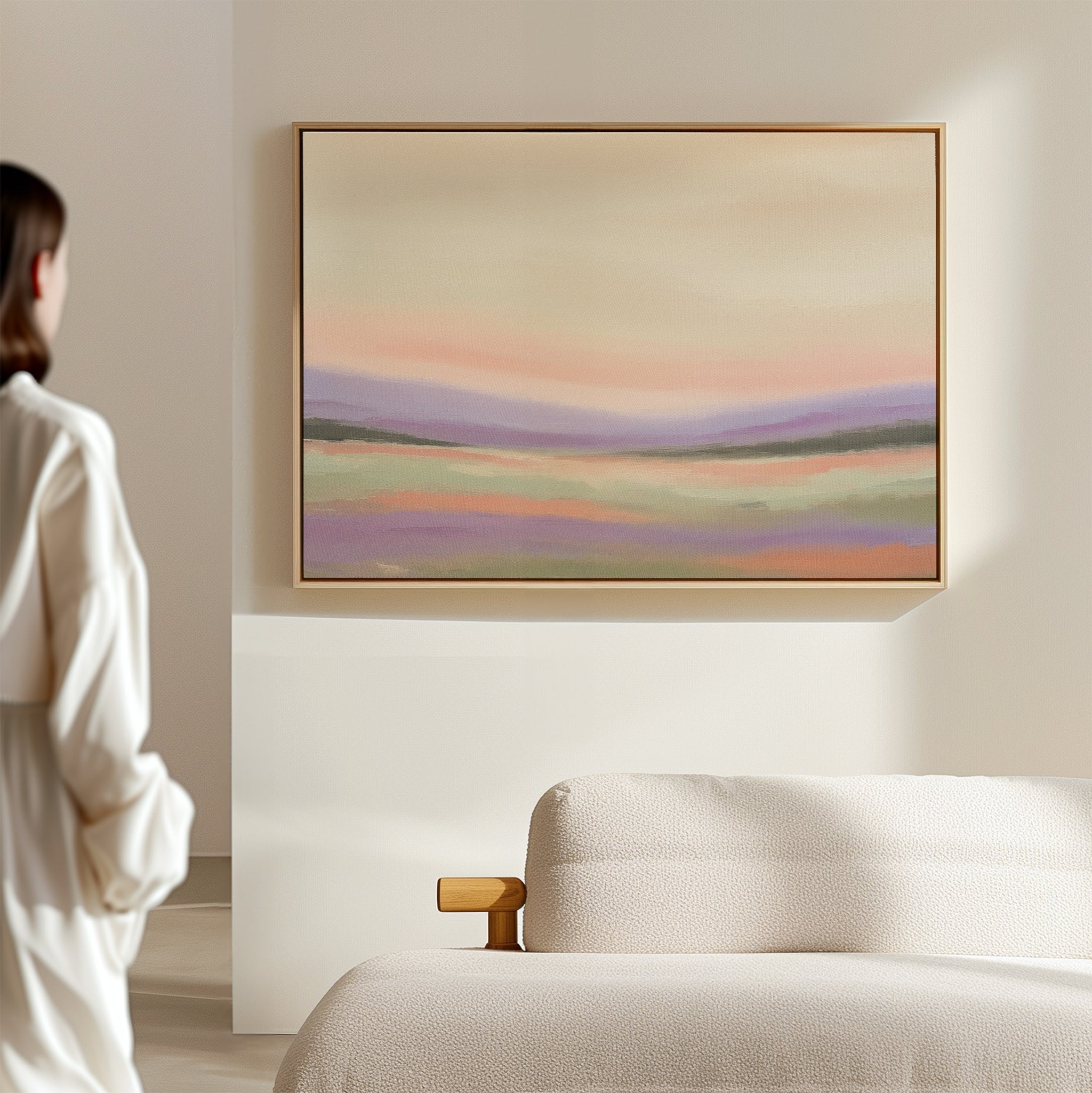 A person in a white robe stands in a minimalist room, gazing at the Lavender Valley Canvas Art - Horizontal, a pastel abstract landscape painting, displayed on a beige wall above a light-colored sofa.