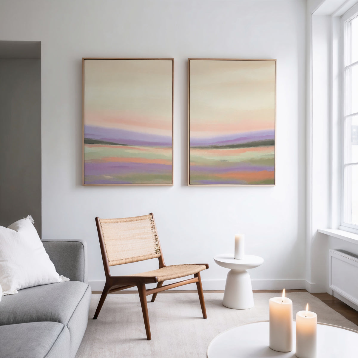 A modern, minimal living room featuring a neutral sofa, woven chair, and the "Lavender Valley - Set Of 2" wall art—pastel landscape prints. A round side table and lit candles add warmth, while large windows invite natural light.