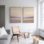 A modern, minimal living room featuring a neutral sofa, woven chair, and the "Lavender Valley - Set Of 2" wall art—pastel landscape prints. A round side table and lit candles add warmth, while large windows invite natural light.