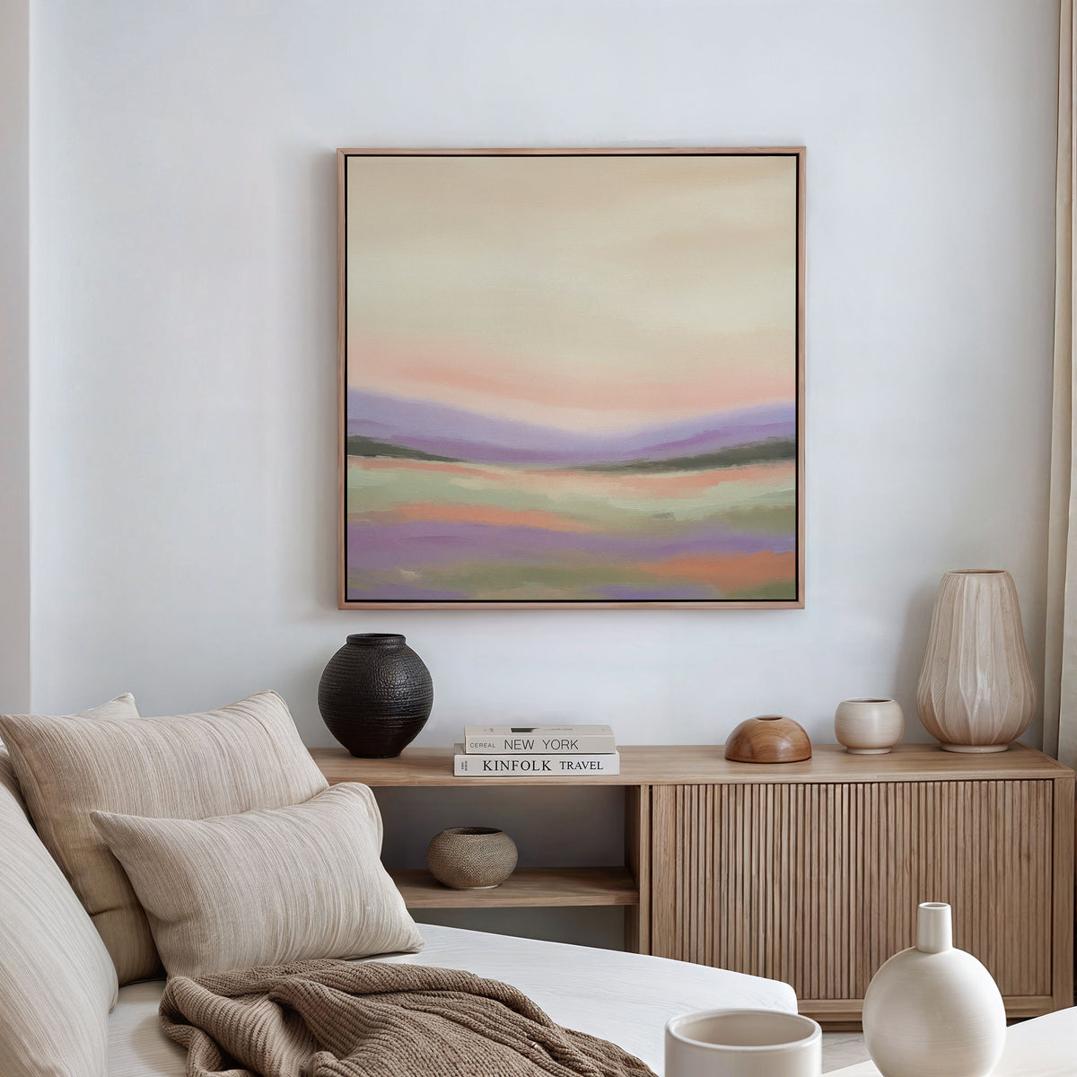 A minimalist living room with a beige sofa, neutral cushions, a wooden sideboard topped with vases and books, and serene decor, featuring the Lavender Valley Canvas Art - Square displayed on a white wall.