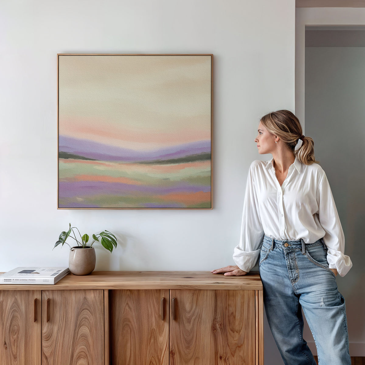 A woman in a white blouse and jeans leans against a wooden cabinet, admiring the Lavender Valley Canvas Art - Square on the wall. A small plant and a book sit atop the cabinet, completing the calm, minimalist decor.