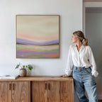 A woman in a white blouse and jeans leans against a wooden cabinet, admiring the Lavender Valley Canvas Art - Square on the wall. A small plant and a book sit atop the cabinet, completing the calm, minimalist decor.