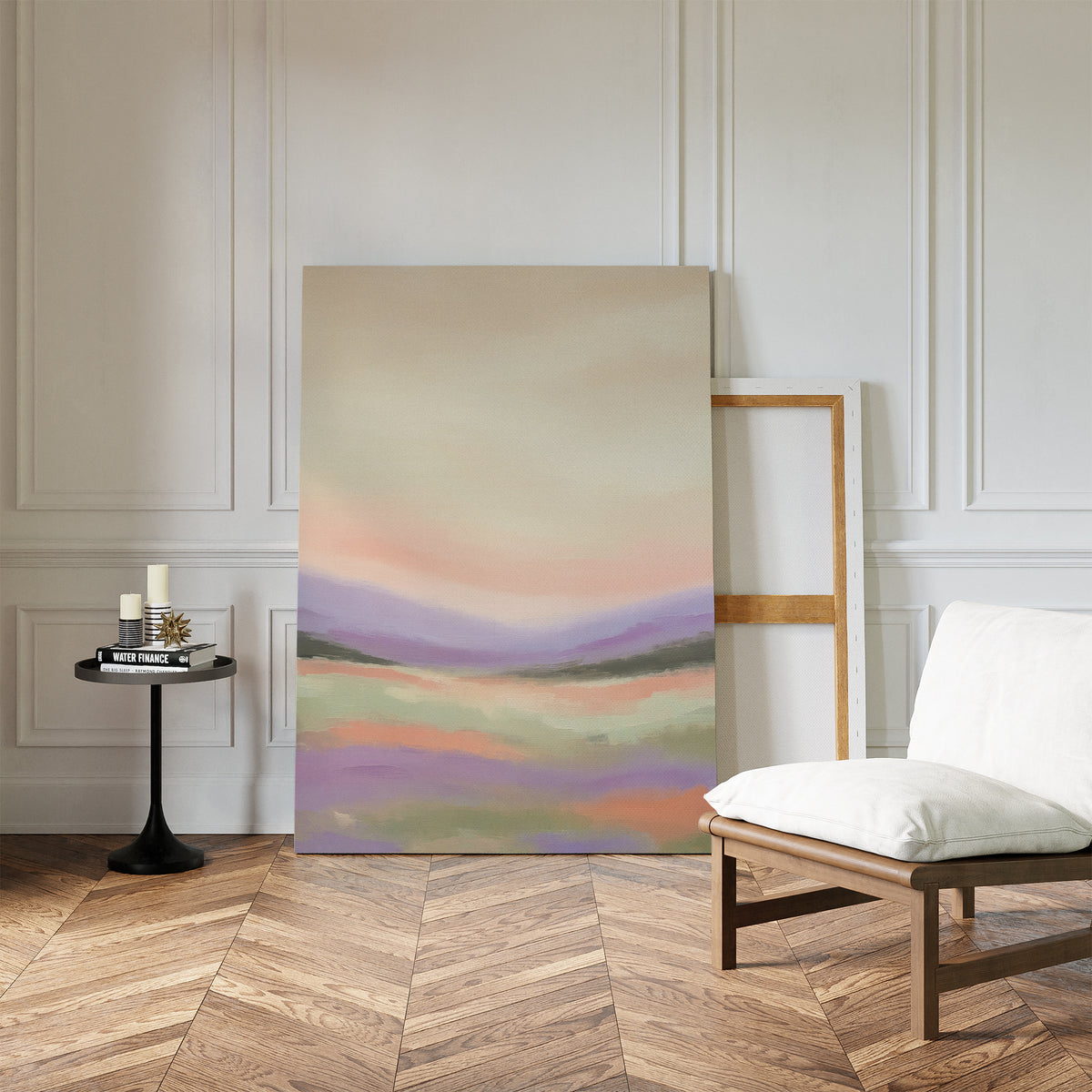 The Lavender Valley Canvas Art - Vertical, a pastel-colored abstract landscape, leans against a white paneled wall in a minimalist room with wooden herringbone floors, a cushioned chair, and a small black round side table with candles and books.