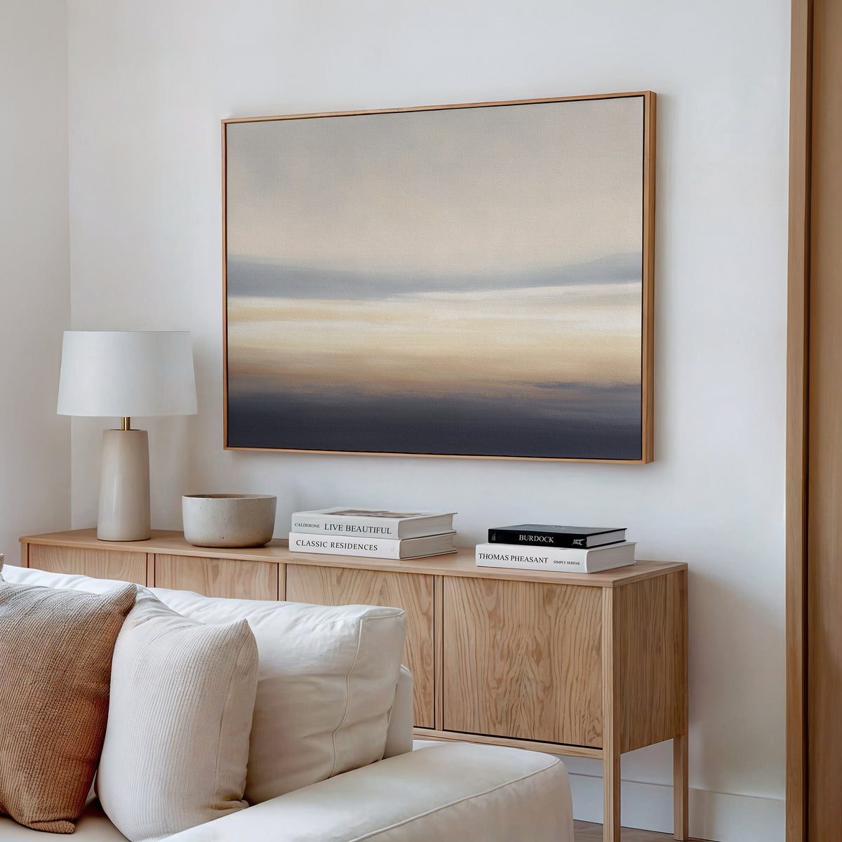 A minimalist living room features a light wooden sideboard, a white lamp, decorative bowl, stacked books, and the Layered Dusk Canvas Art - Horizontal from Nordic Park in soft neutral tones hanging above.