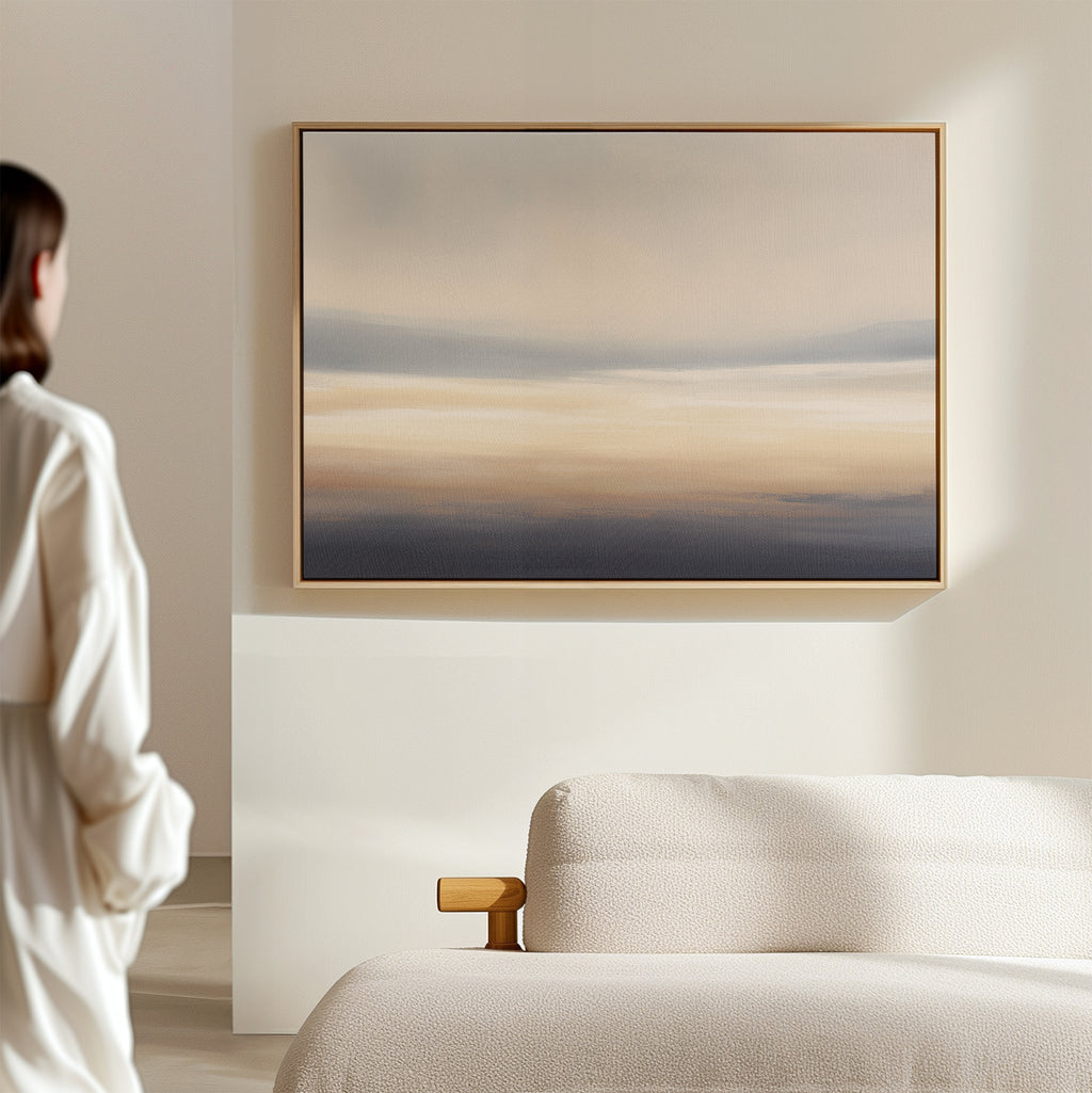 In a minimalist room, a person admires the Layered Dusk Canvas Art - Horizontal, featuring calming blue, beige, and gray tones inspired by Nordic Park, displayed above a light sofa on a cream-colored wall.