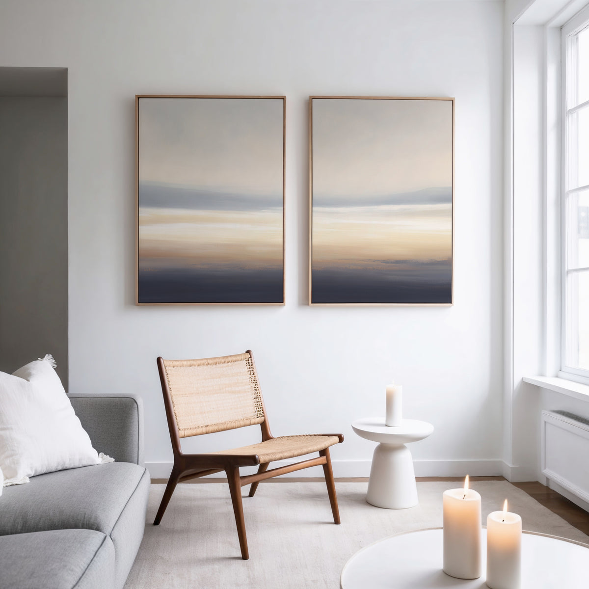 A minimalist living room features a light gray sofa, woven chair, small white side table, and three lit candles. Above the chair hang Layered Dusk - Set Of 2 abstract prints. Natural light pours through large windows.