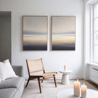 A minimalist living room features a light gray sofa, woven chair, small white side table, and three lit candles. Above the chair hang Layered Dusk - Set Of 2 abstract prints. Natural light pours through large windows.