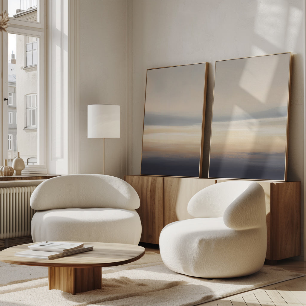 A modern living room featuring Layered Dusk - Set Of 2 rounded white armchairs, a low wooden coffee table, sideboard, abstract wall art, floor lamp, and large windows creates a serene atmosphere filled with natural light.