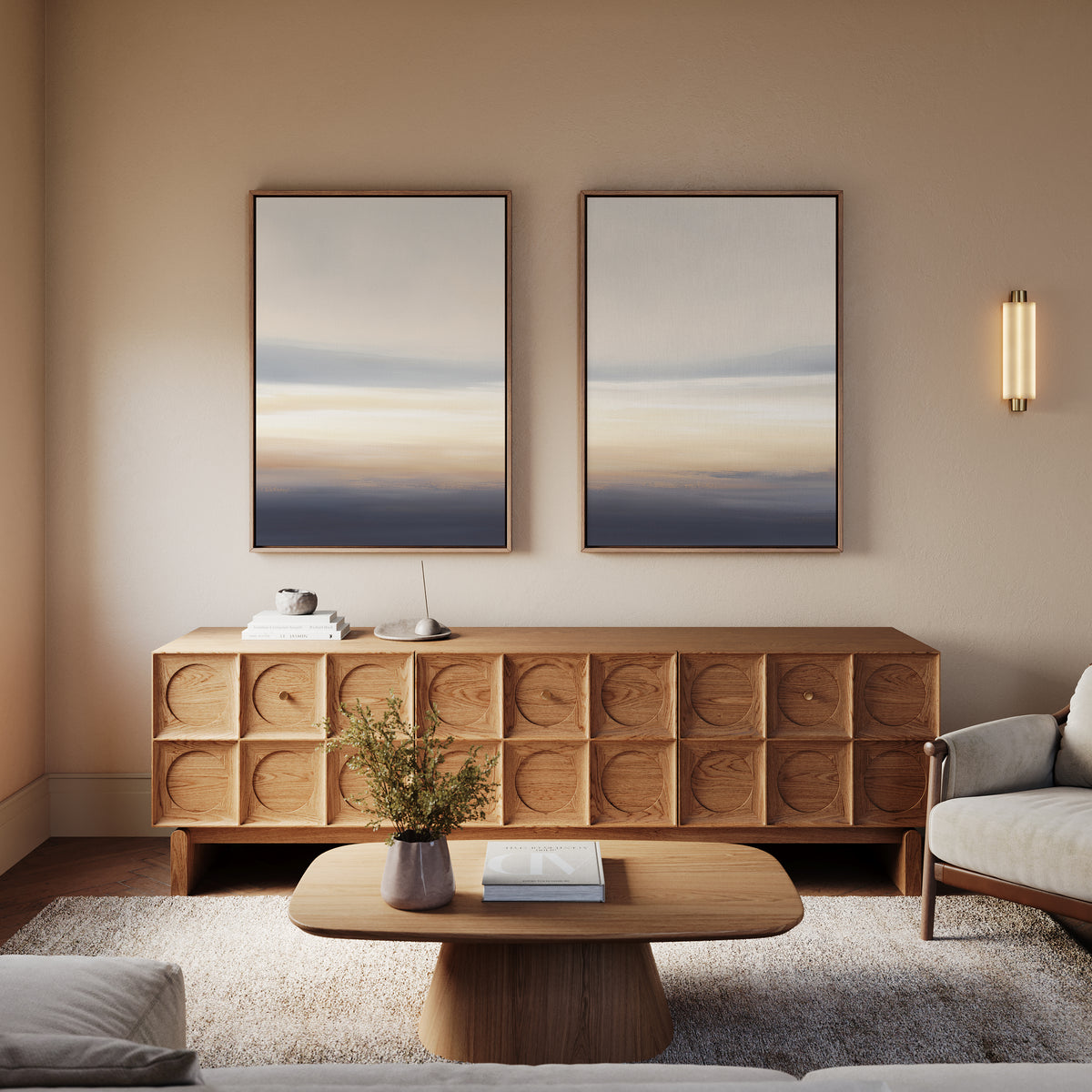 A modern living room features a wooden sideboard, matching coffee table, beige armchair, and Layered Dusk - Set Of 2 abstract wall art; a small vase with flowers on the table enhances the room’s contemporary charm.