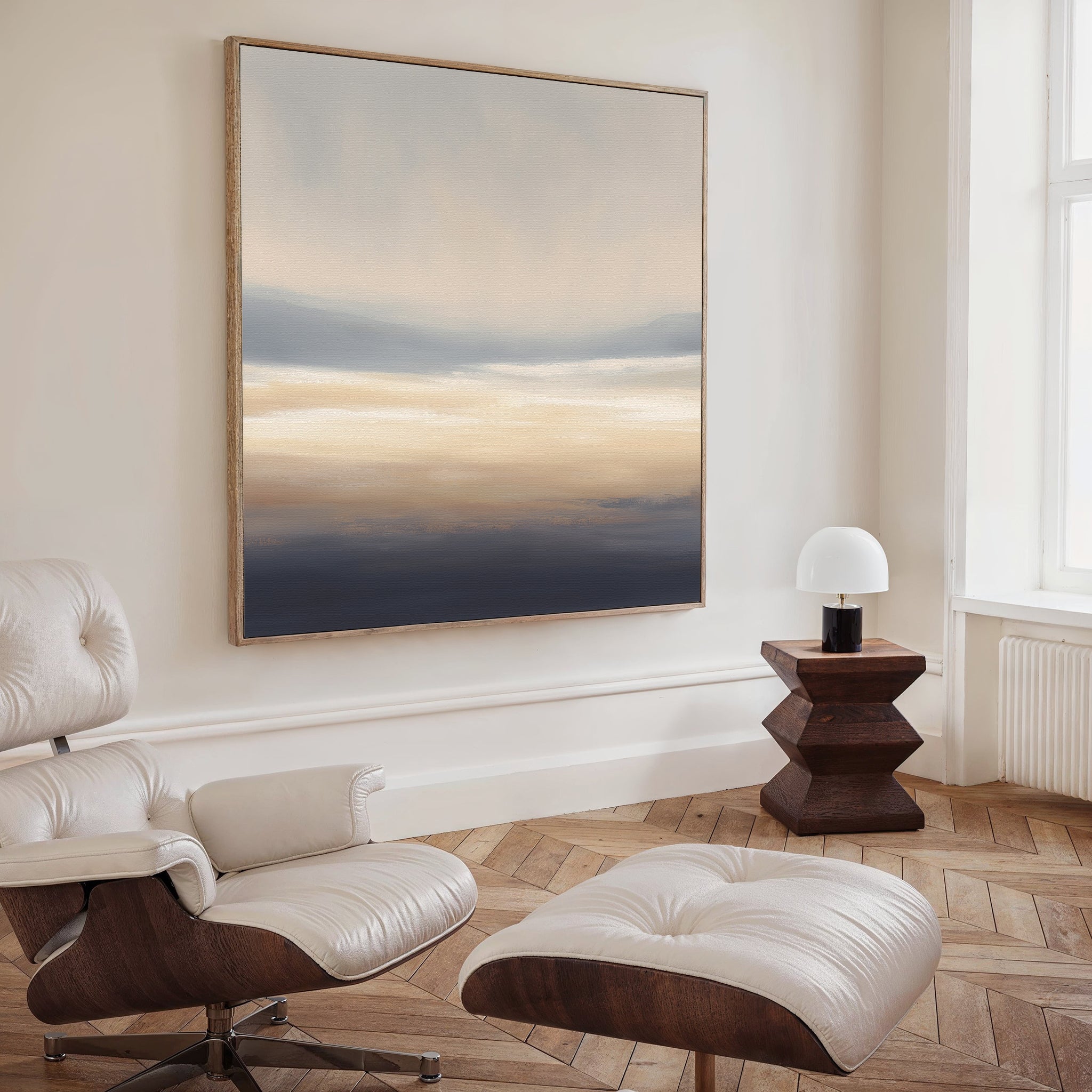 A modern living room with a white leather chair and ottoman, dark wood side table with a round lamp, and the Layered Dusk Canvas Art - Square in soft beige, blue, and gray tones brings tranquility to the space.