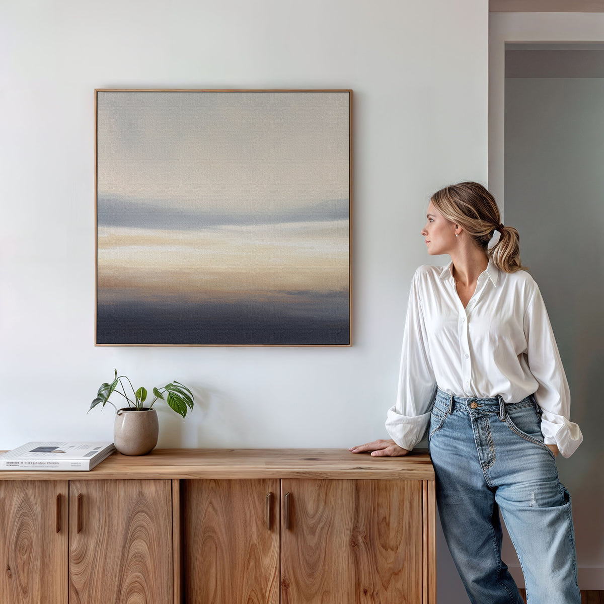 A woman in a white blouse and jeans leans against a wooden cabinet, gazing at the Layered Dusk Canvas Art - Square. A potted plant and a closed book sit on the cabinet beneath the abstract wall art.