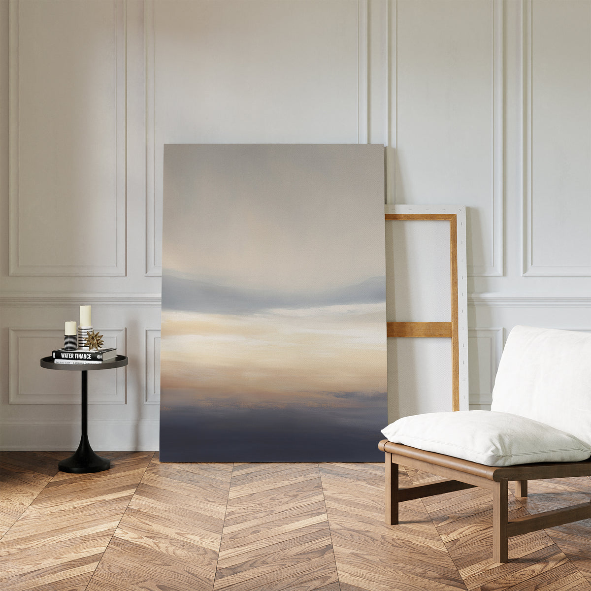 A minimalist room with herringbone wood floors, a white cushioned chair, a round black side table with candles and books, and the Layered Dusk Canvas Art - Vertical as wall art, leaning against a white paneled wall.