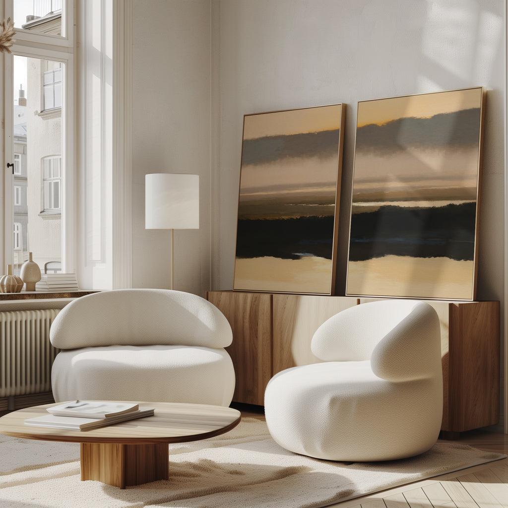 A modern living room features two rounded white armchairs, a low wooden coffee table, and the Layered Valley Dawn - Set Of 2 wall art on a wooden sideboard. Large windows brighten the serene, neutral decor with natural light.
