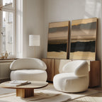 A modern living room features two rounded white armchairs, a low wooden coffee table, and the Layered Valley Dawn - Set Of 2 wall art on a wooden sideboard. Large windows brighten the serene, neutral decor with natural light.