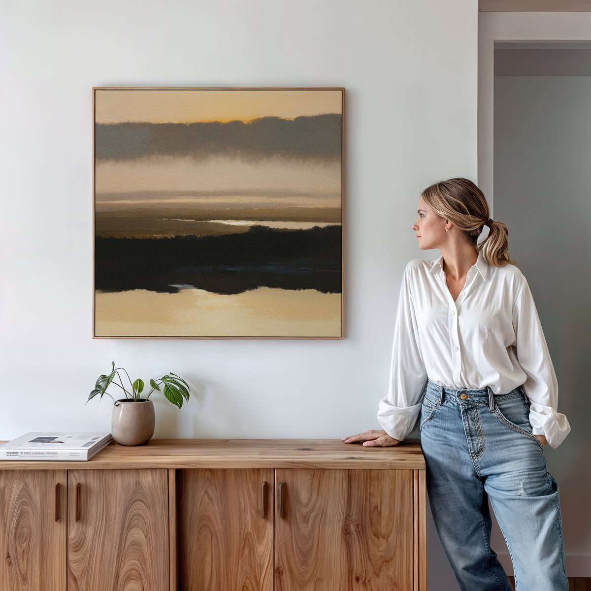 A woman in a white shirt and jeans stands beside a wooden sideboard, admiring the Layered Valley Dawn Canvas Art - Square, a tranquil landscape piece hanging on the wall. A small plant and a book sit on the sideboard.