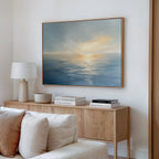 A minimalist living room features a wooden sideboard with books, lamp, and decor. Above, the Luminous Calm Canvas Art - Horizontal showcases a tranquil ocean at sunset. A cream sofa in front adds to the serene coastal vibe.