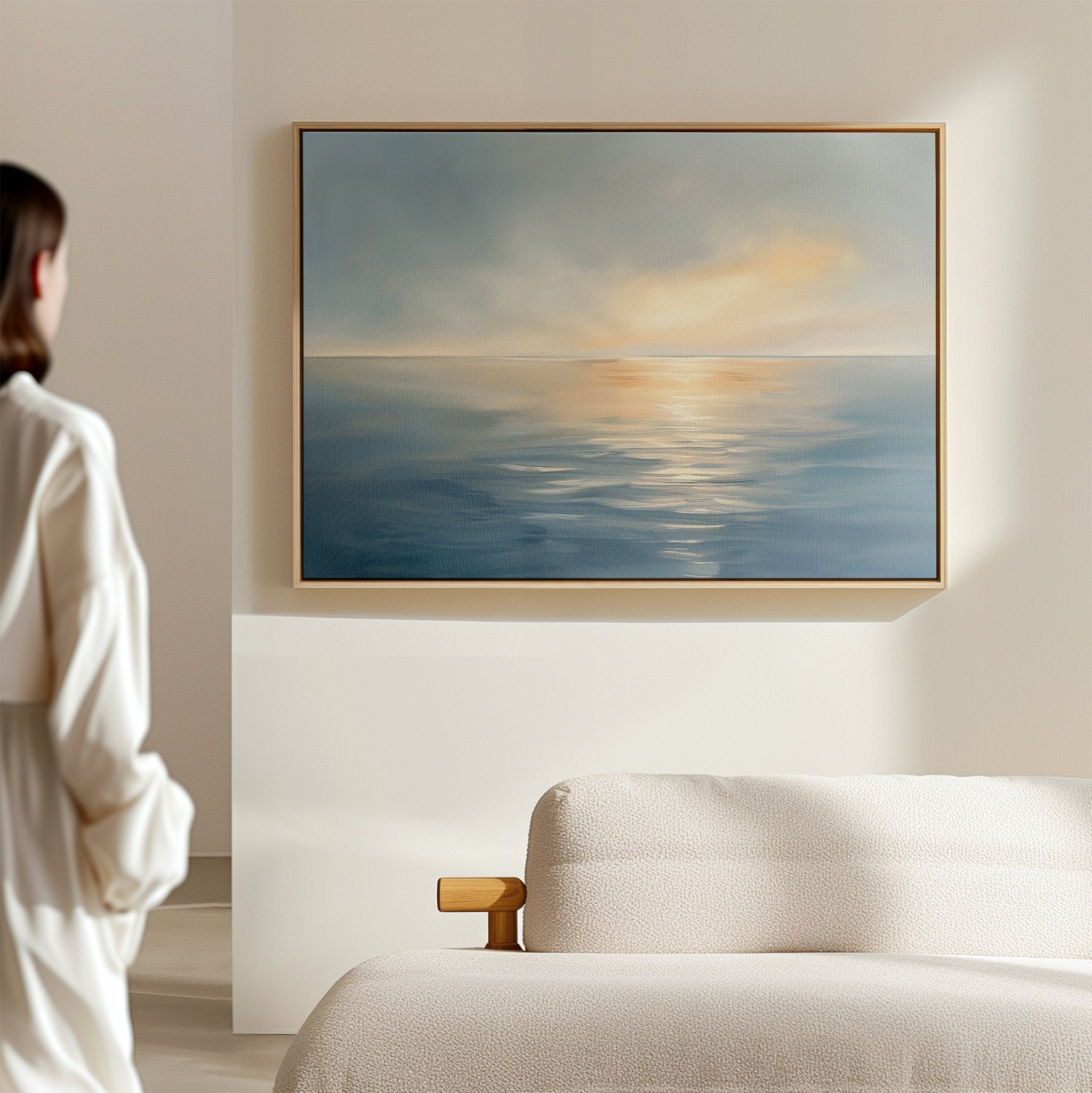 A person stands in a softly lit room, admiring the "Luminous Calm Canvas Art - Horizontal," its serene ocean sunset tones enhancing the tranquil ambiance above a beige sofa.