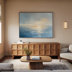 A modern living room with a wooden coffee table, sideboard, beige sofa, textured rug, and a vase of flowers. The Luminous Calm Canvas Art - Horizontal above the sideboard and warm lighting evoke a serene coastal vibe.
