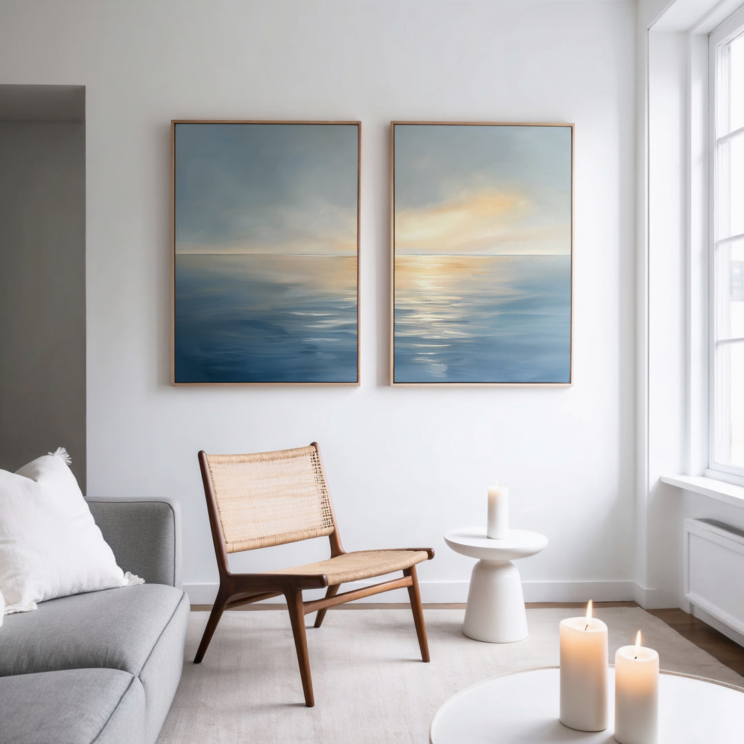 A minimal, modern living room features the Luminous Calm - Set of 2 canvas prints of a tranquil sea at sunset above a wooden chair and round table with candles, white pillows, and soft lighting for a serene coastal vibe.