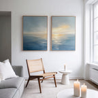 A minimal, modern living room features the Luminous Calm - Set of 2 canvas prints of a tranquil sea at sunset above a wooden chair and round table with candles, white pillows, and soft lighting for a serene coastal vibe.