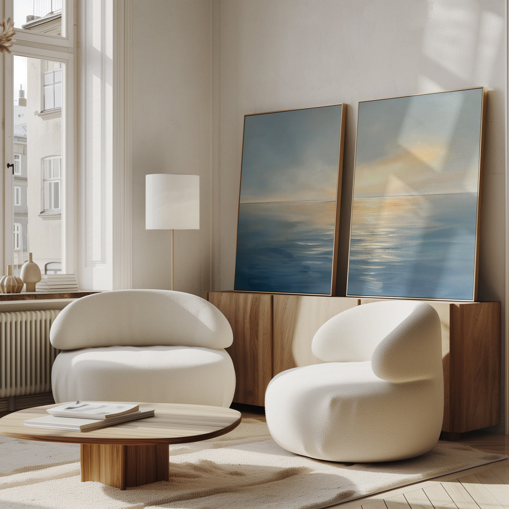 A bright, modern living room features two white curved armchairs, a round wooden coffee table, a wooden sideboard, and "Luminous Calm - Set of 2" canvas prints in coastal hues. Natural light enhances the serene atmosphere.