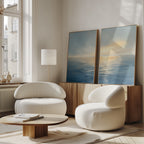 A bright, modern living room features two white curved armchairs, a round wooden coffee table, a wooden sideboard, and "Luminous Calm - Set of 2" canvas prints in coastal hues. Natural light enhances the serene atmosphere.