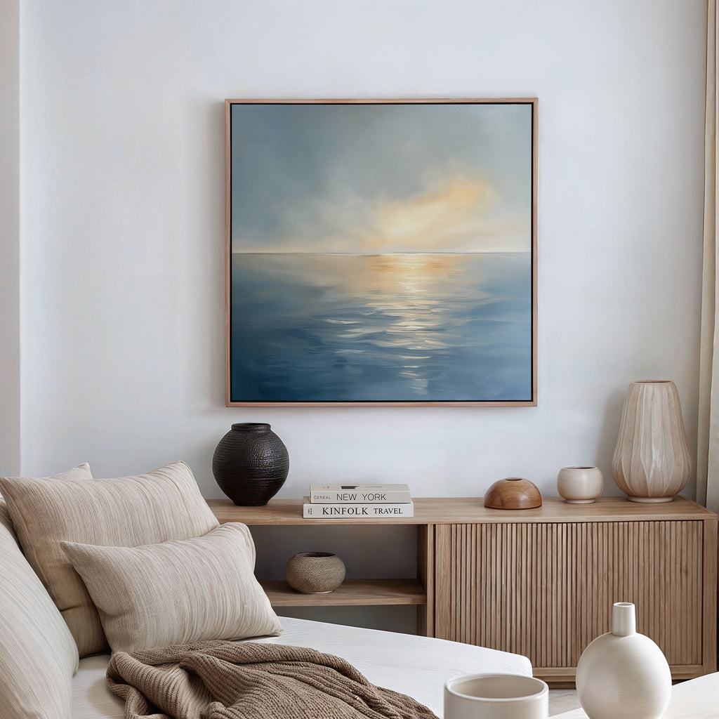 A cozy living room with a beige sofa, neutral cushions, wooden sideboard with books and vases, and the Luminous Calm Canvas Art - Square depicting a serene sea and sky in soothing coastal tones hanging on the wall.
