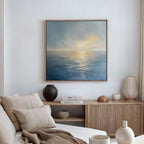 A cozy living room with a beige sofa, neutral cushions, wooden sideboard with books and vases, and the Luminous Calm Canvas Art - Square depicting a serene sea and sky in soothing coastal tones hanging on the wall.