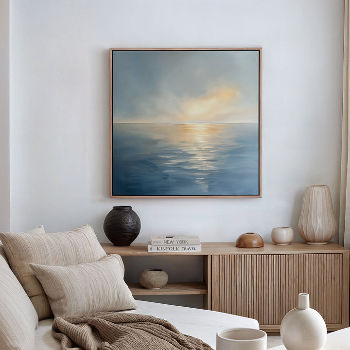 A cozy living room with a beige sofa, neutral cushions, wooden sideboard with books and vases, and the Luminous Calm Canvas Art - Square depicting a serene sea and sky in soothing coastal tones hanging on the wall.