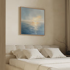 A minimalist bedroom features a beige upholstered bed with white bedding and soft pillows. The Luminous Calm Canvas Art - Square hangs above, infusing the softly lit space with a coastal vibe. A bare branch decorates the bedside table.