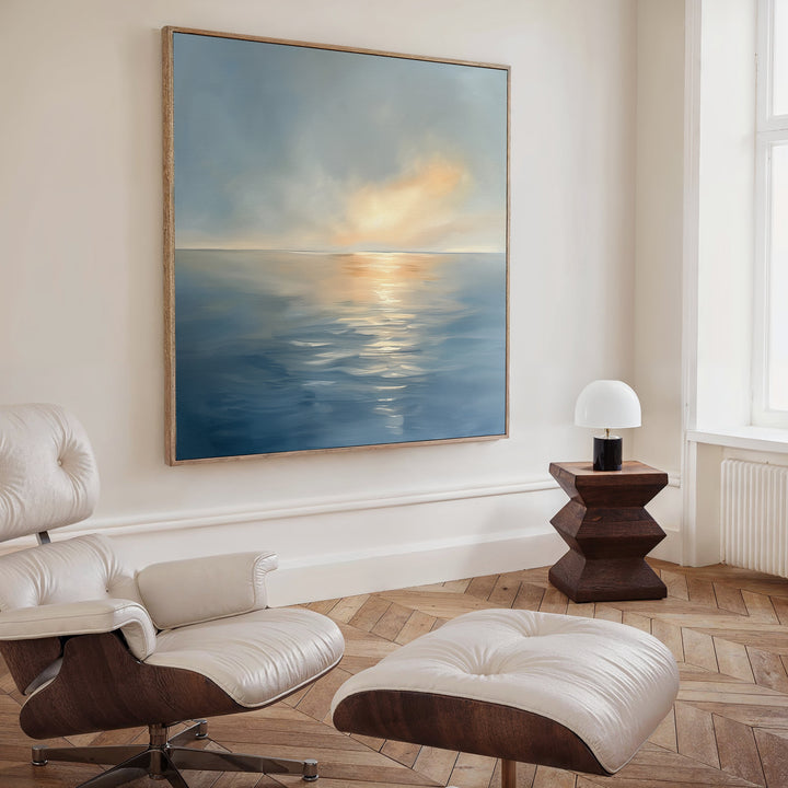 A modern living room featuring a white leather lounge chair and ottoman, a wooden side table with a white lamp, and the Luminous Calm Canvas Art - Square showcasing a serene sea at sunset hanging on the wall.