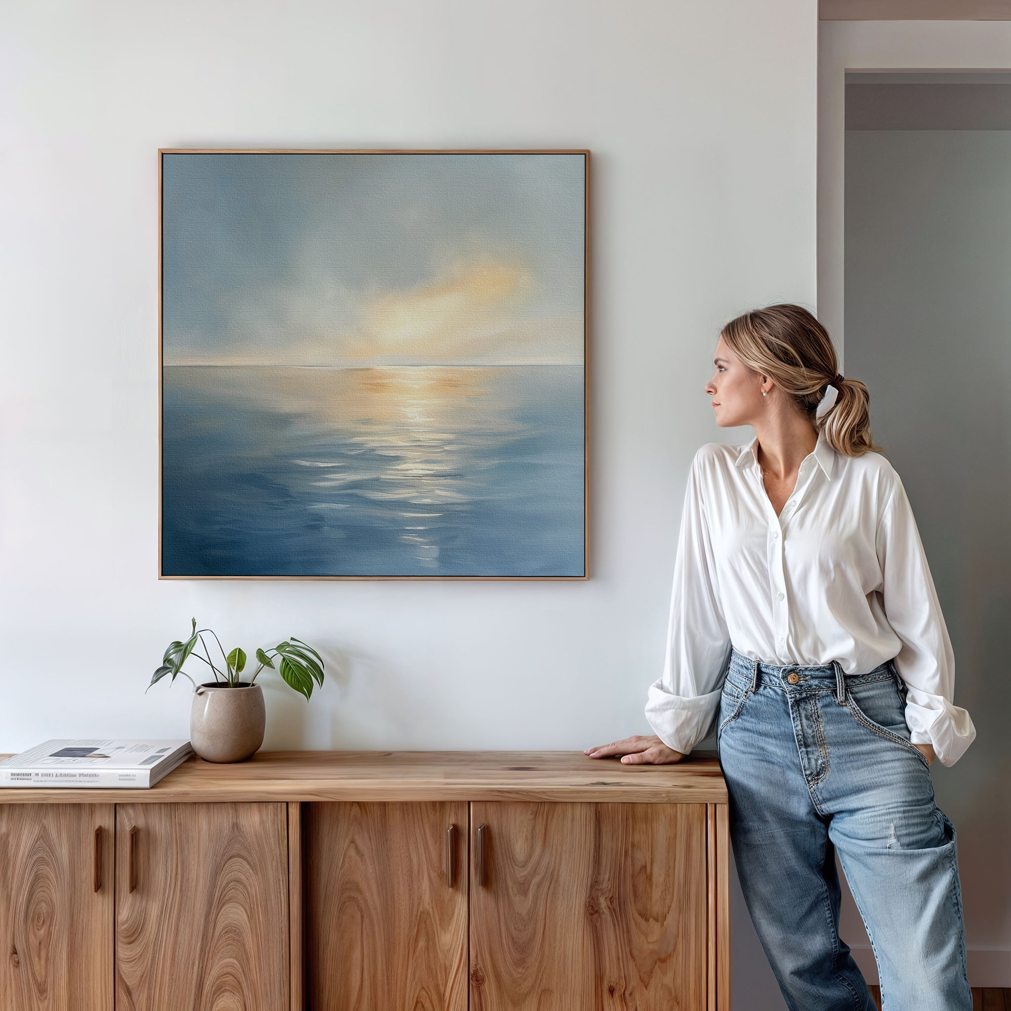 A woman in a white blouse and jeans stands by a wooden cabinet, admiring the Luminous Calm Canvas Art - Square, which features a serene ocean sunset. A small plant and books on the cabinet enhance the tranquil display.