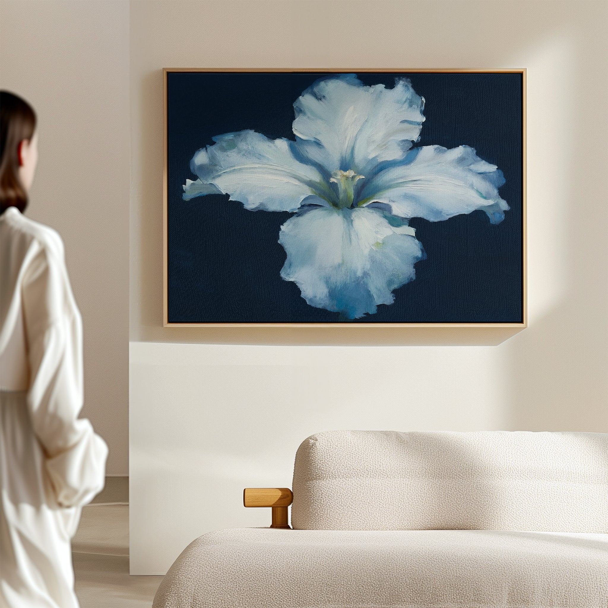 A person in a white shirt admires the Luminous Iris Canvas Art - Horizontal, featuring glowing white and blue petals against a dark background, displayed above a light-colored sofa on a beige wall.