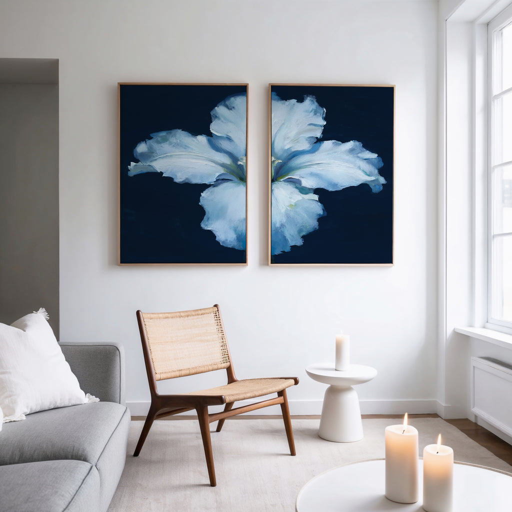 A modern living room features a light gray sofa, woven chair, and round white side table with candles. Two Luminous Iris - Set of 2 canvas prints with blue backgrounds hang above the chair as natural light streams through tall windows.
